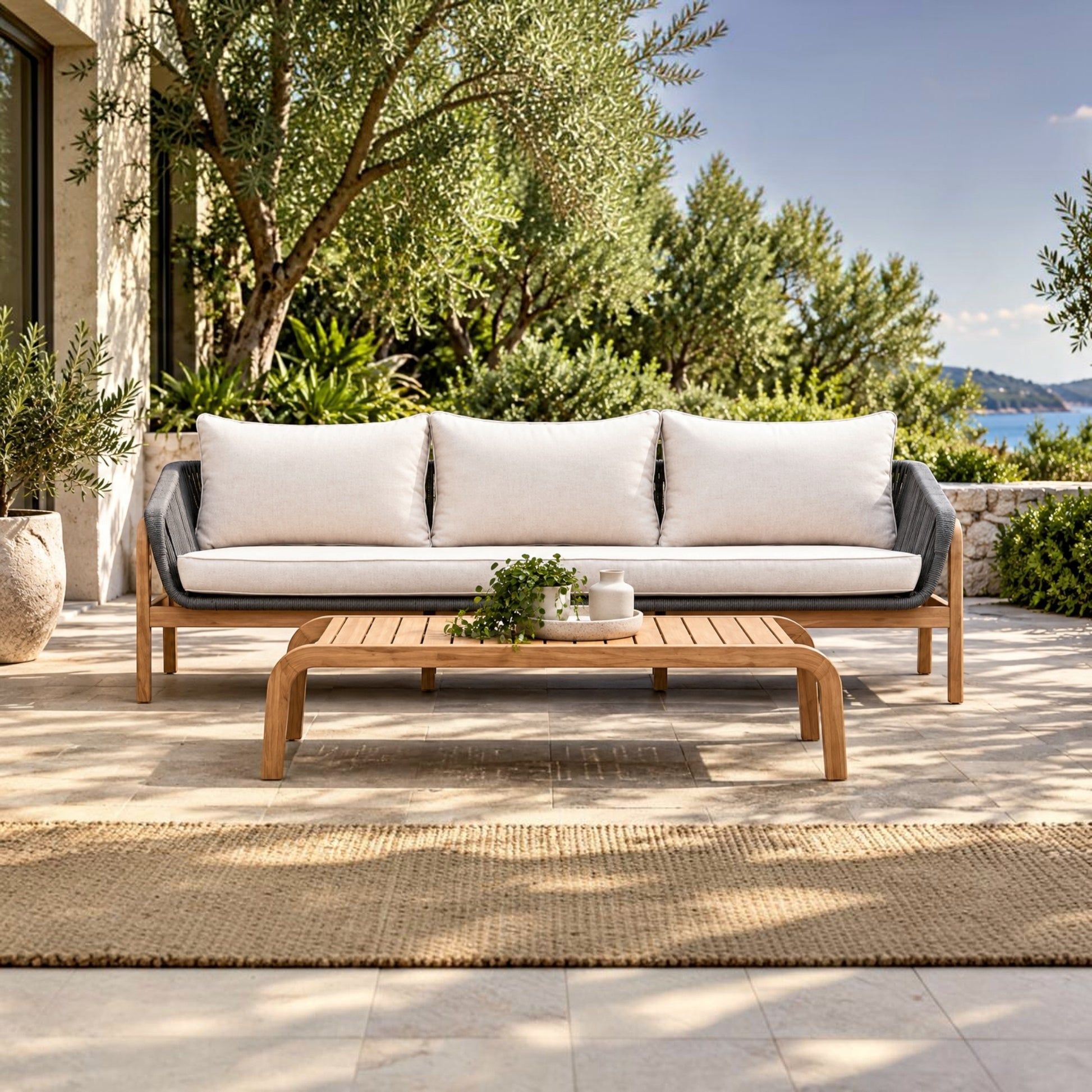 A modern outdoor patio sofa with white cushions and a light wood frame, paired with a matching wooden coffee table on a tiled patio with a woven rug, olive trees, and a scenic ocean view in the background.