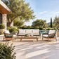 A 5-piece outdoor patio furniture set including a three-seater sofa, two armchairs, and a rectangular coffee table, arranged on a woven rug in a luxurious backyard setting with a pool, trees, and a stone house.