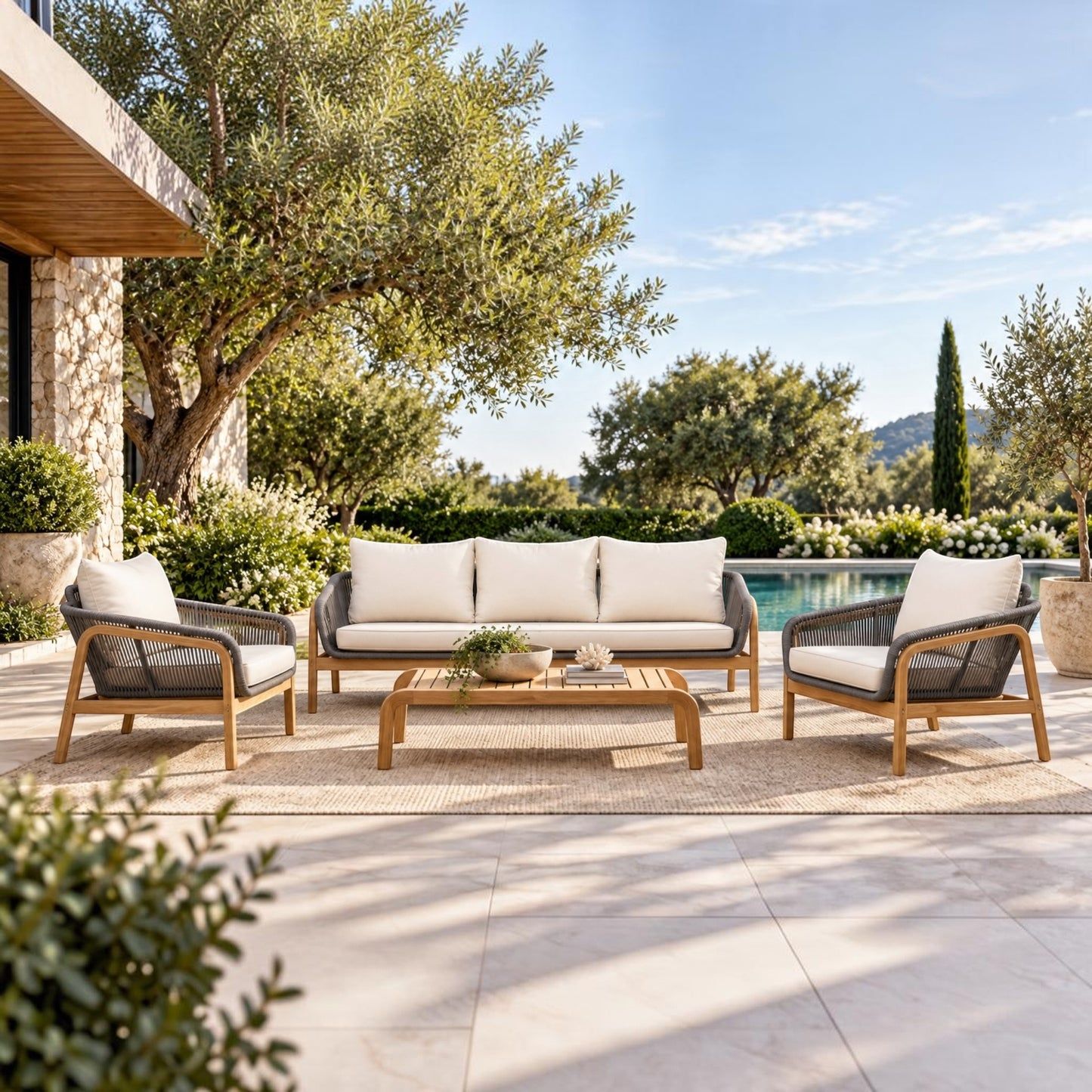 A 5-piece outdoor patio furniture set including a three-seater sofa, two armchairs, and a rectangular coffee table, arranged on a woven rug in a luxurious backyard setting with a pool, trees, and a stone house.