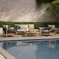 Outdoor patio furniture set by a swimming pool, including a three-seat sofa, two armchairs, and a low rectangular coffee table, arranged on a stone deck against a hedge and textured wall.