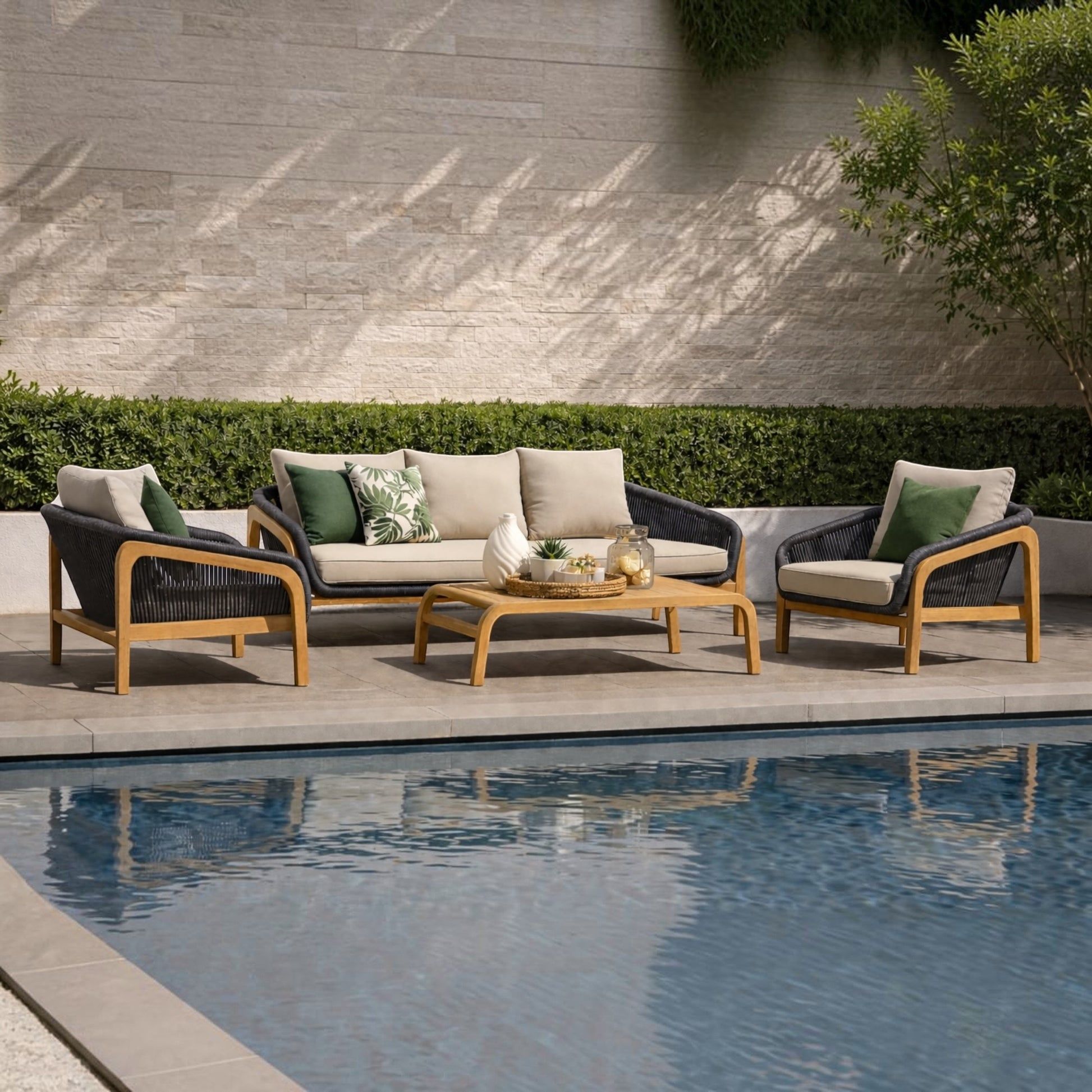 Outdoor patio furniture set by a swimming pool, including a three-seat sofa, two armchairs, and a low rectangular coffee table, arranged on a stone deck against a hedge and textured wall.