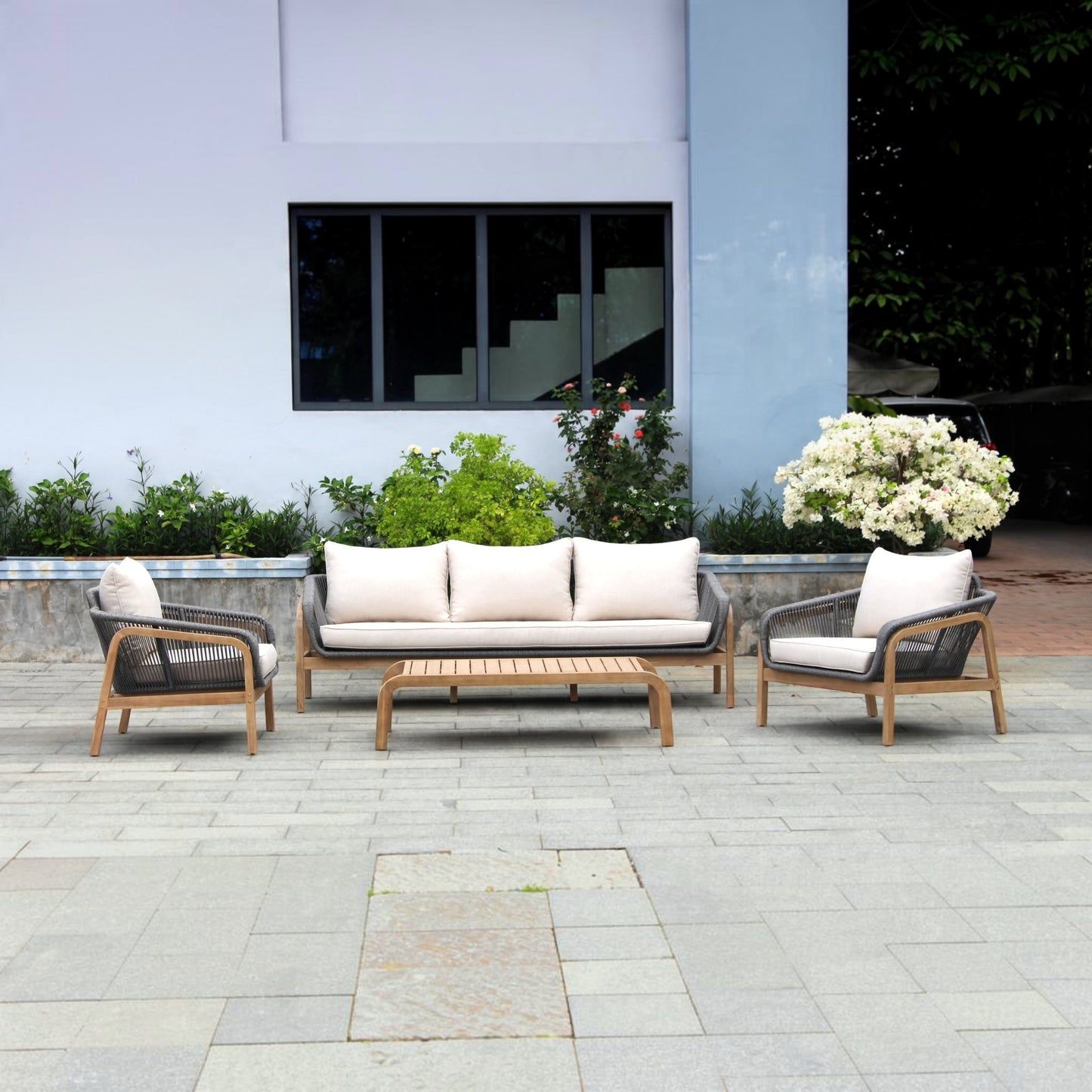 Outdoor patio furniture set including a three-seat sofa, two armchairs, and a rectangular slat coffee table, arranged on a stone-paved terrace in front of a modern building.