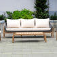 Outdoor wooden three-seater sofa with light beige cushions and a matching slatted coffee table on a paved patio with green plants in the background.