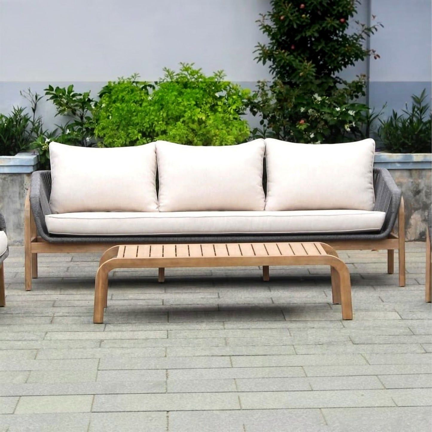 Outdoor wooden three-seater sofa with light beige cushions and a matching slatted coffee table on a paved patio with green plants in the background.