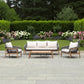 Outdoor patio furniture set including a three-seat sofa, two armchairs, and a rectangular coffee table, arranged on a stone patio in a landscaped garden with green hedges and flowers. The furniture has light wood frames, dark woven seats, and off-white cushions.