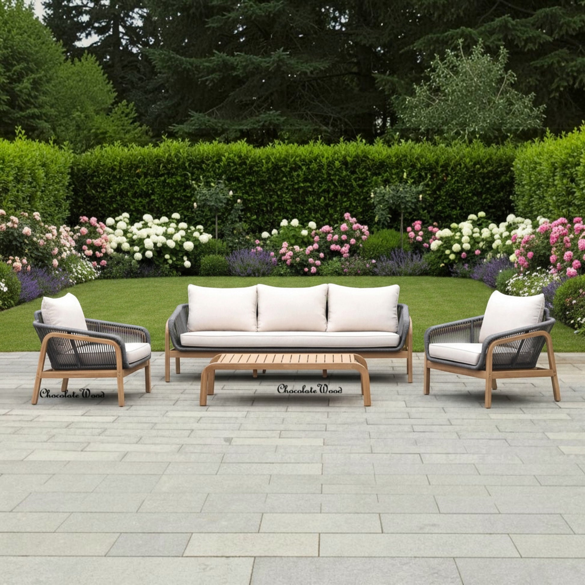 Outdoor patio furniture set including a three-seat sofa, two armchairs, and a rectangular coffee table, arranged on a stone patio in a landscaped garden with green hedges and flowers. The furniture has light wood frames, dark woven seats, and off-white cushions.