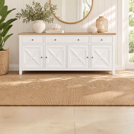 A white wooden sideboard cabinet with four doors featuring an X-brace design, set in a bright room with a light wood top, styled with a vase of greenery, a mirror, and a woven rug.