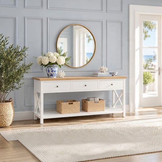 A white console table with a light wood top is styled in a bright entryway with a round gold-framed mirror, blue and white ceramic vase, white flowers, and two woven storage baskets on the shelf.