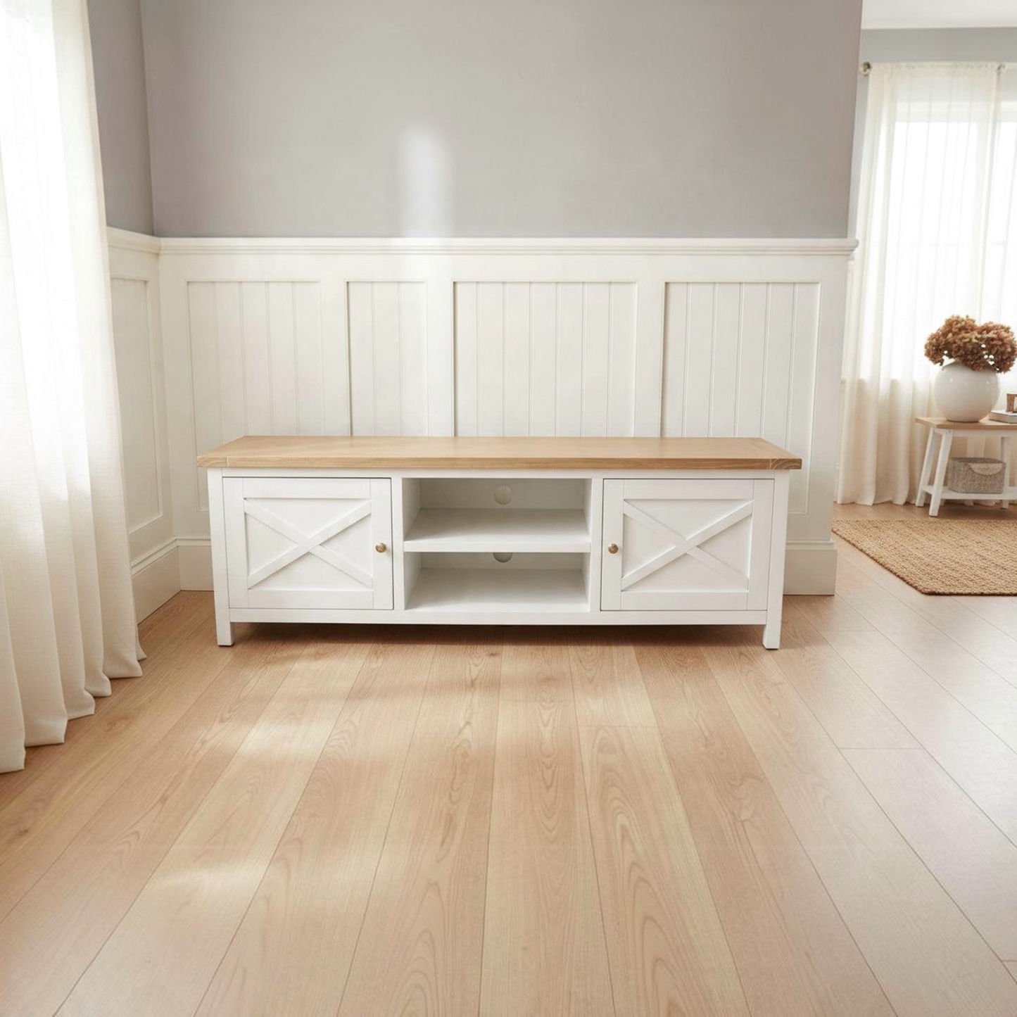 A white wooden media console with a light natural wood top, shown in a bright, modern room with light wood flooring and white wainscoting on the wall.