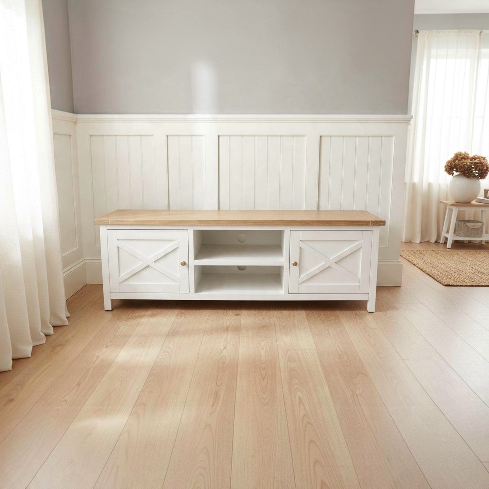 A white wooden media console with a light natural wood top, shown in a bright, modern room with light wood flooring and white wainscoting on the wall.