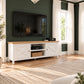 A white wooden TV stand with a light oak top and brass hardware, styled in a living room against a dark green shiplap wall with a mounted television.