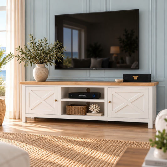 A white wooden TV stand with a light oak top, styled in a bright living room under a wall-mounted television. The stand features two cabinet doors with an X-pattern, an open center shelf with a basket and decorative items, and small brass knobs.