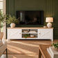 A white wooden TV stand with a light oak top, featuring two side cabinets with cross-shaped panels and an open center shelf, styled in a living room against a dark green paneled wall.