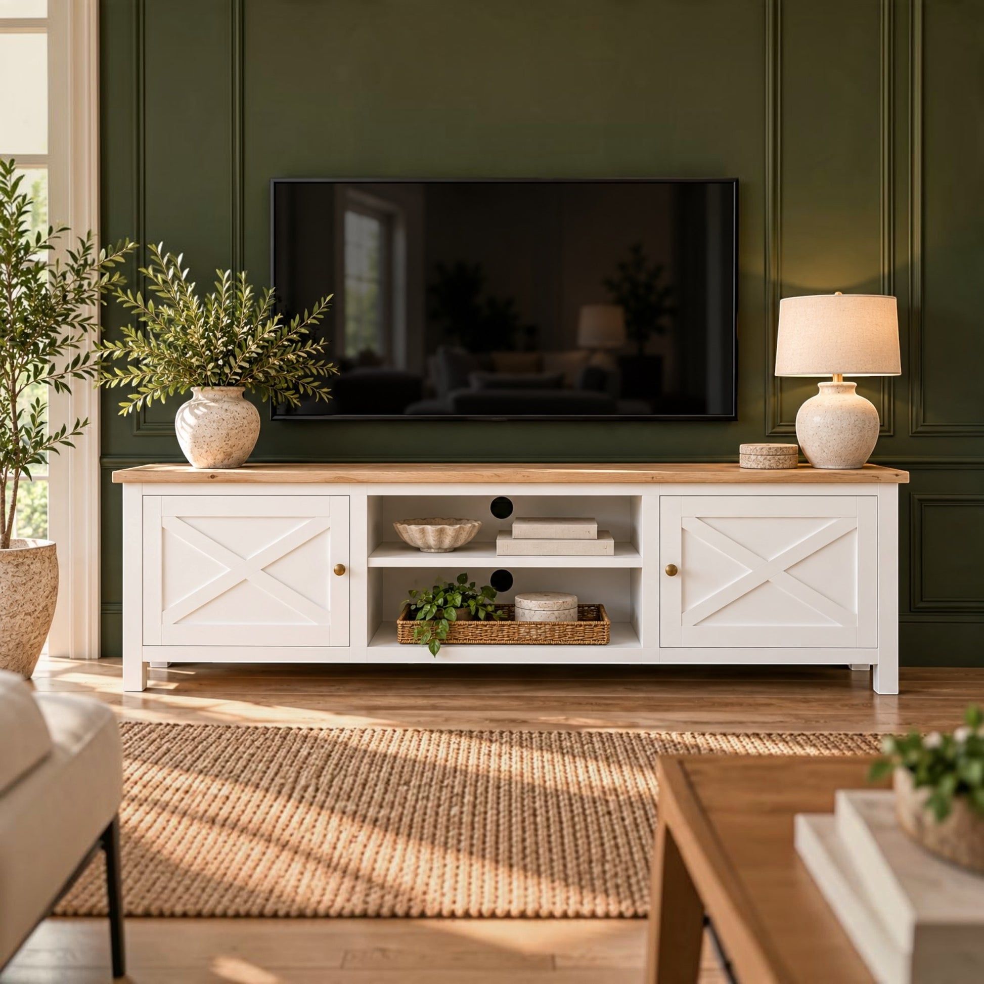 A white wooden TV stand with a light oak top, featuring two side cabinets with cross-shaped panels and an open center shelf, styled in a living room against a dark green paneled wall.