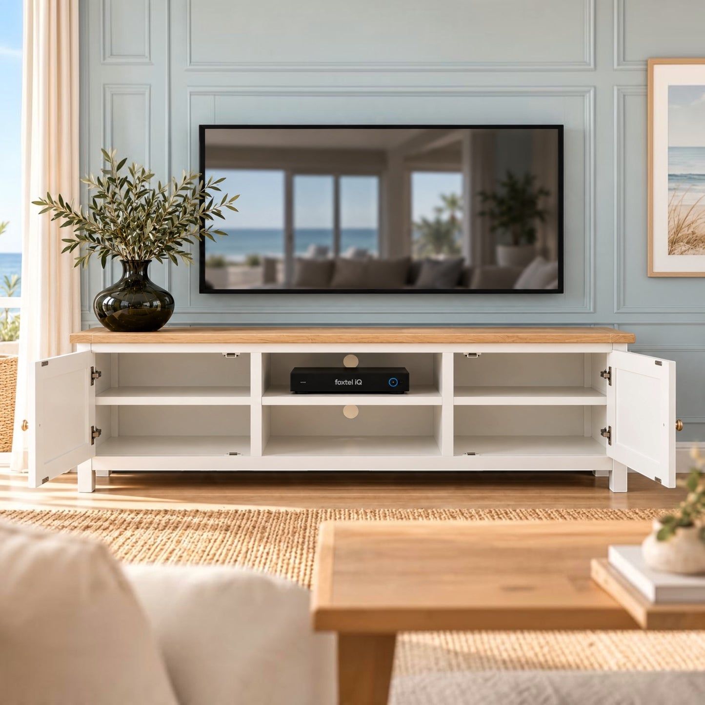 A white TV stand with a light wood top, shown in a styled living room setting with the doors open to display storage compartments. A television, a vase of greenery, and a woven rug are visible in the background.