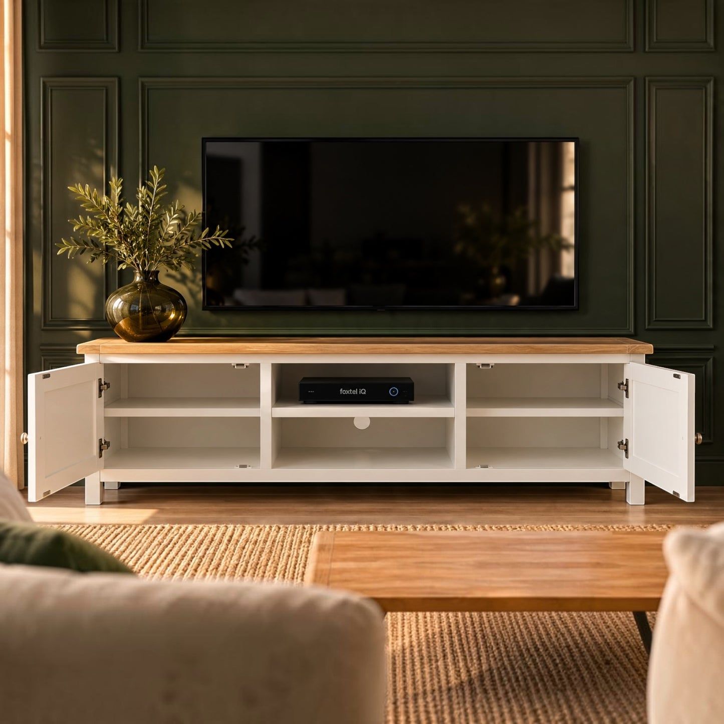 A white TV stand with a light wood top and open doors, styled in a modern living room setting with a television on top and a plant in a vase on the left.