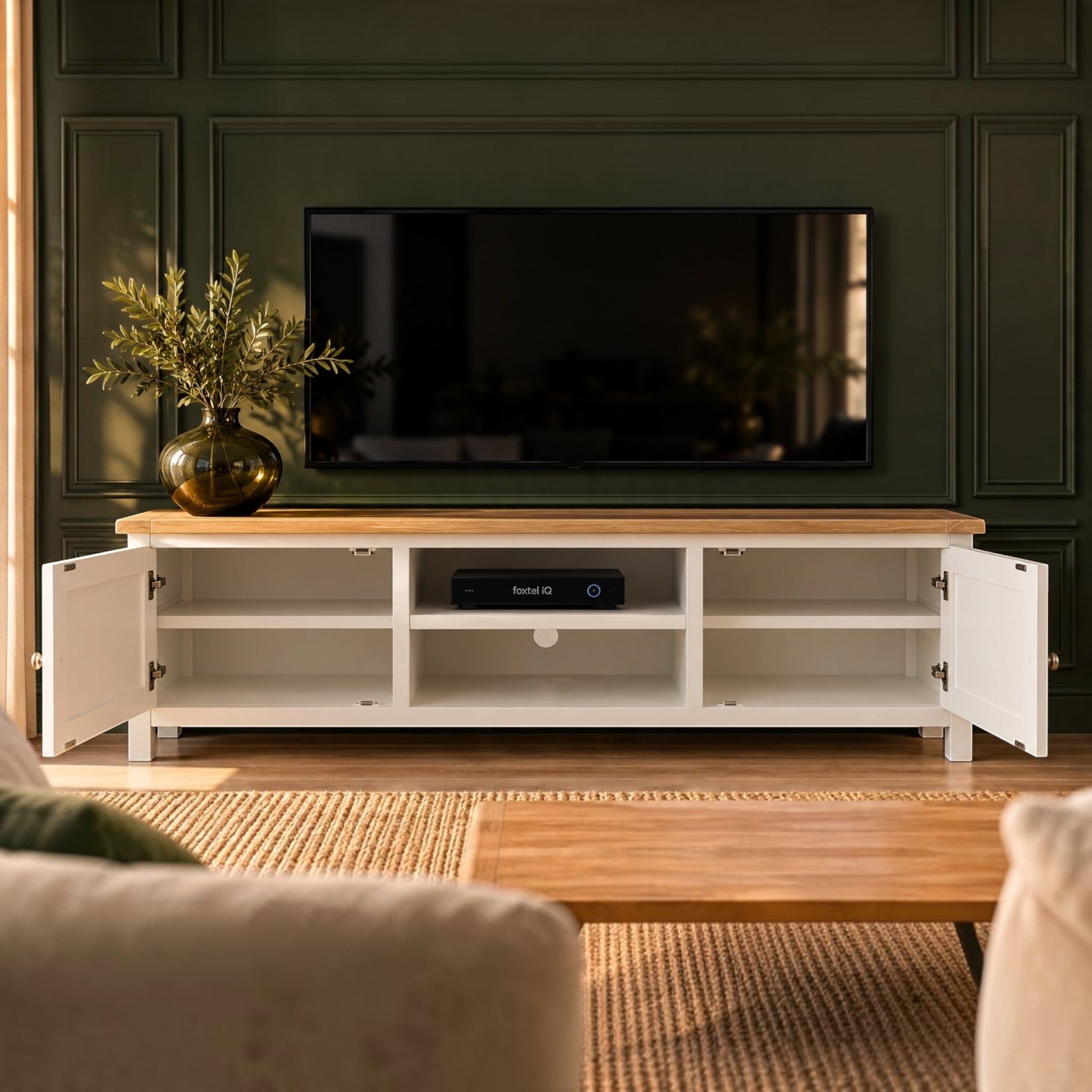 A white TV stand with a light wood top and open doors, styled in a modern living room setting with a television on top and a plant in a vase on the left.