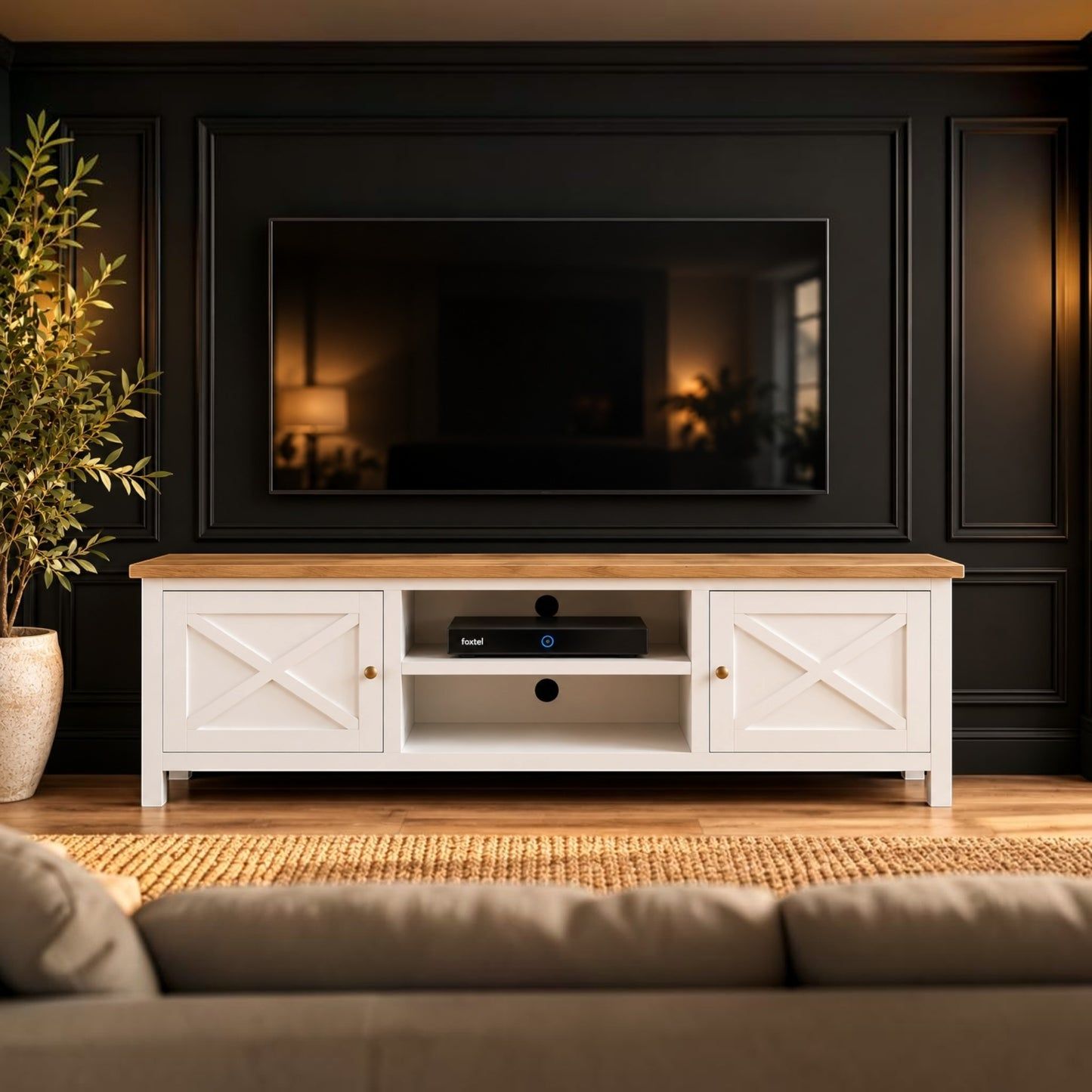 A white TV stand with a light wood top and decorative barn-style doors, positioned in a living room setting in front of a black paneled wall with a television.