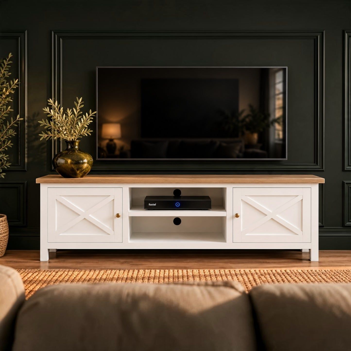 A white TV stand with a light wood top and brass hardware, styled in a living room setting with a television, plants, and a sofa against a dark paneled wall.