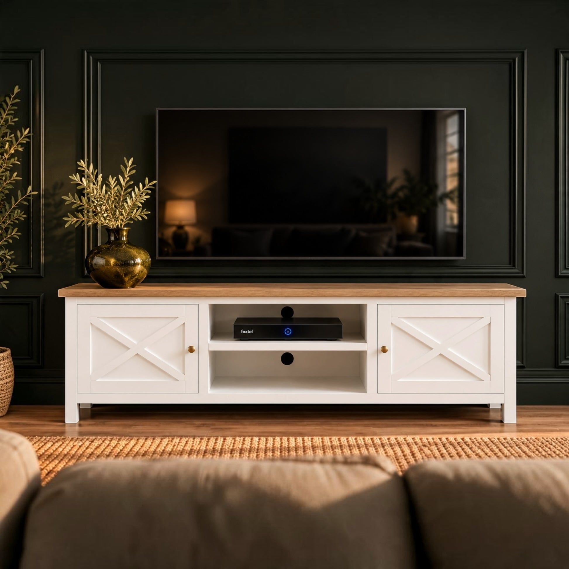A white TV stand with a light wood top and brass hardware, styled in a living room setting with a television, plants, and a sofa against a dark paneled wall.