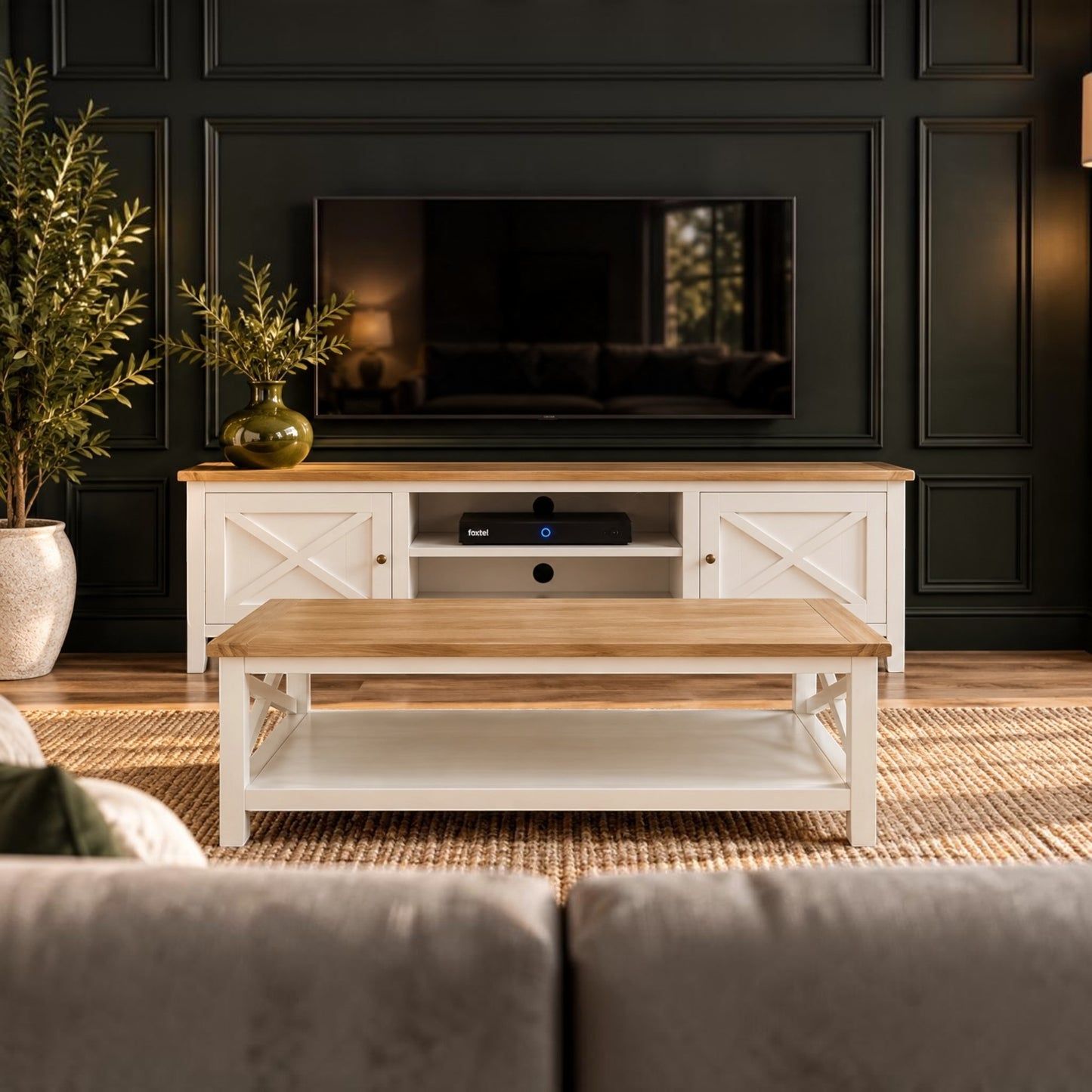 A white TV stand with a light oak wood top and matching coffee table in a styled living room with a dark paneled wall and a television.