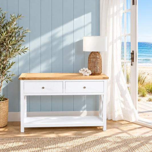 A white wooden console table with a natural wood top, two drawers, and an open lower shelf, styled in a bright beach-themed room with a blue paneled wall, ocean view, and white curtains.