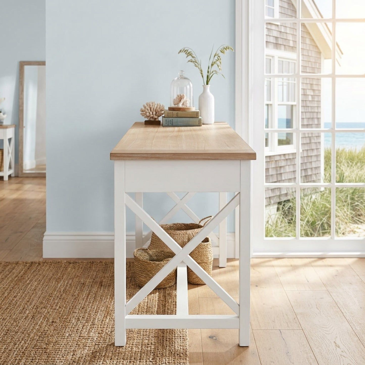 A white wooden writing desk with a light natural wood top and an X-shaped support frame on the legs, styled in a bright coastal room with a window looking out to the ocean.