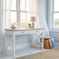 A white writing desk with a light wood top and X-frame legs, styled in a bright, coastal-themed room with sheer white curtains, a lamp, and a woven basket.