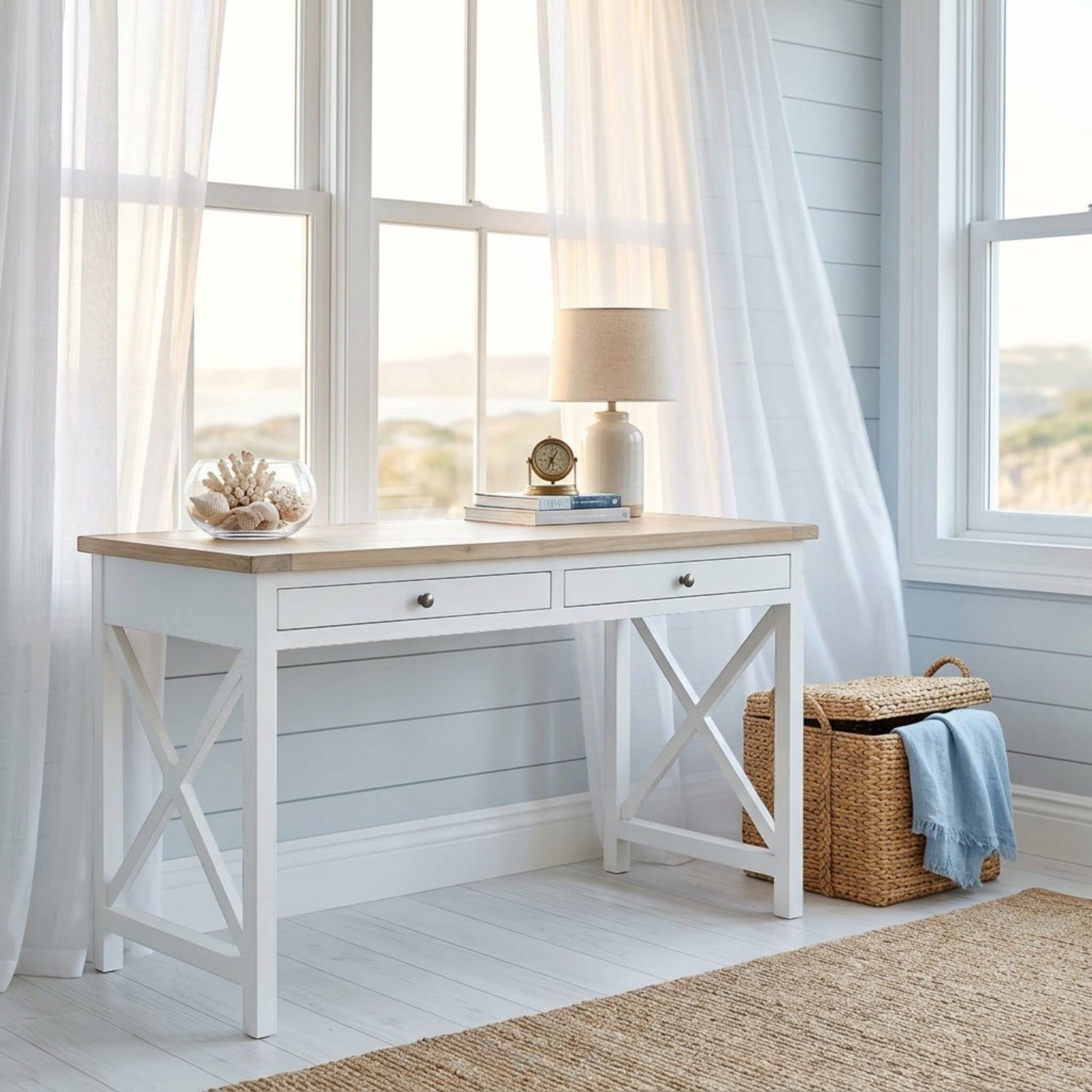 A white writing desk with a light wood top and X-frame legs, styled in a bright, coastal-themed room with sheer white curtains, a lamp, and a woven basket.