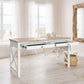 A white writing desk with a light wood top and two open drawers, styled in a bright, coastal-themed room with white curtains, a woven basket, and a driftwood mirror.