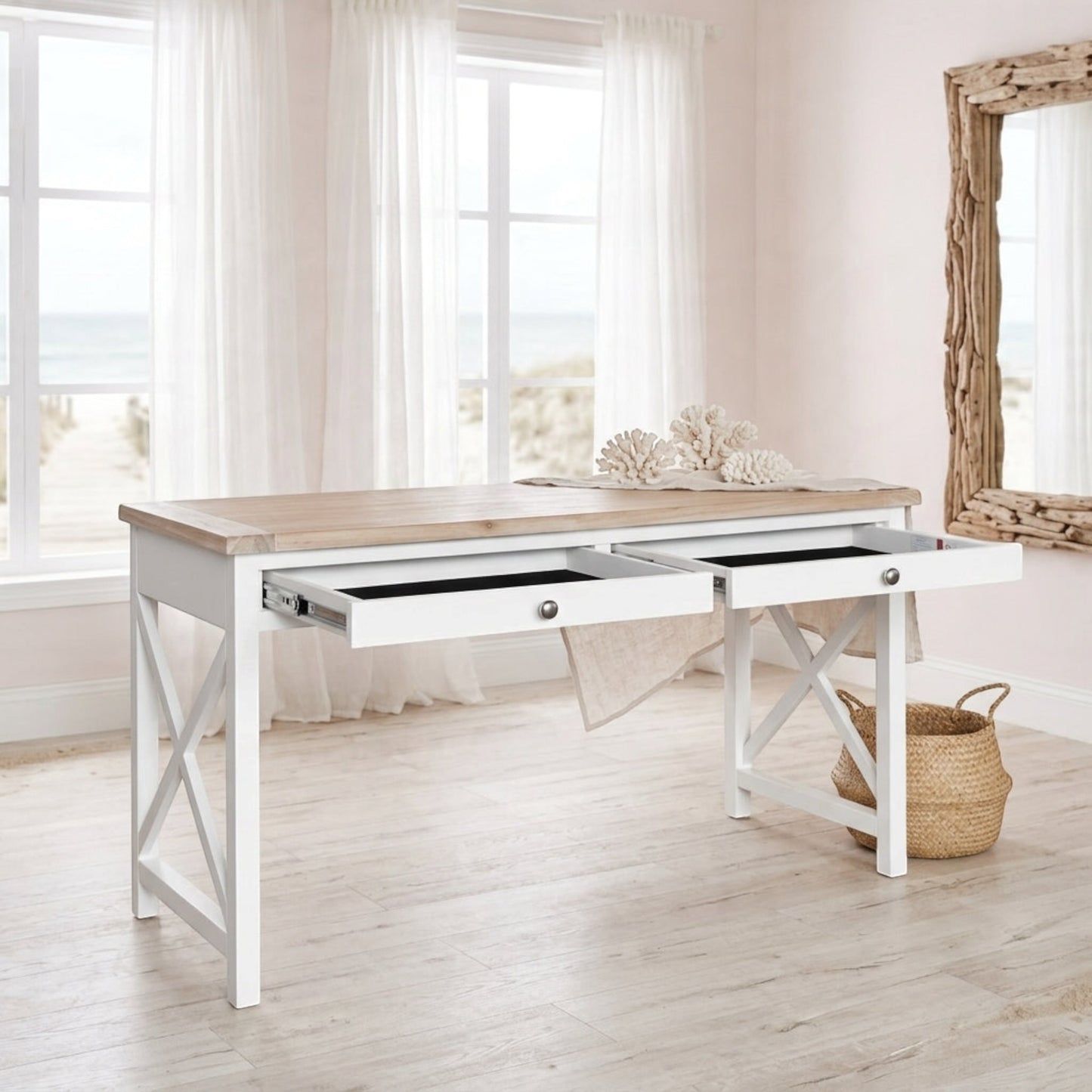 A white writing desk with a light wood top and two open drawers, styled in a bright, coastal-themed room with white curtains, a woven basket, and a driftwood mirror.