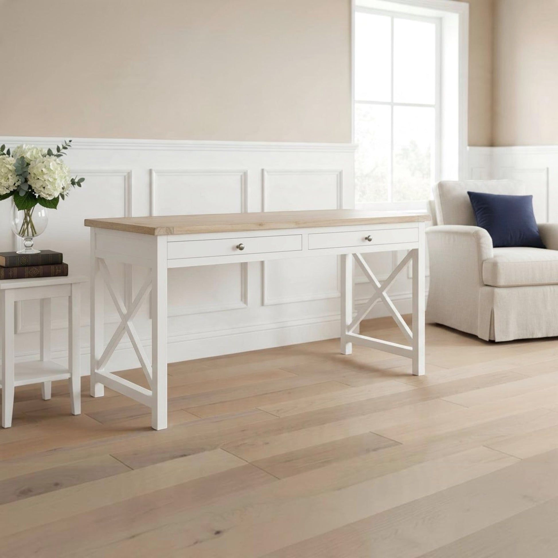 A white writing desk with a light wood top and two drawers, styled in a bright, neutral-toned room next to a small side table with flowers and an armchair.
