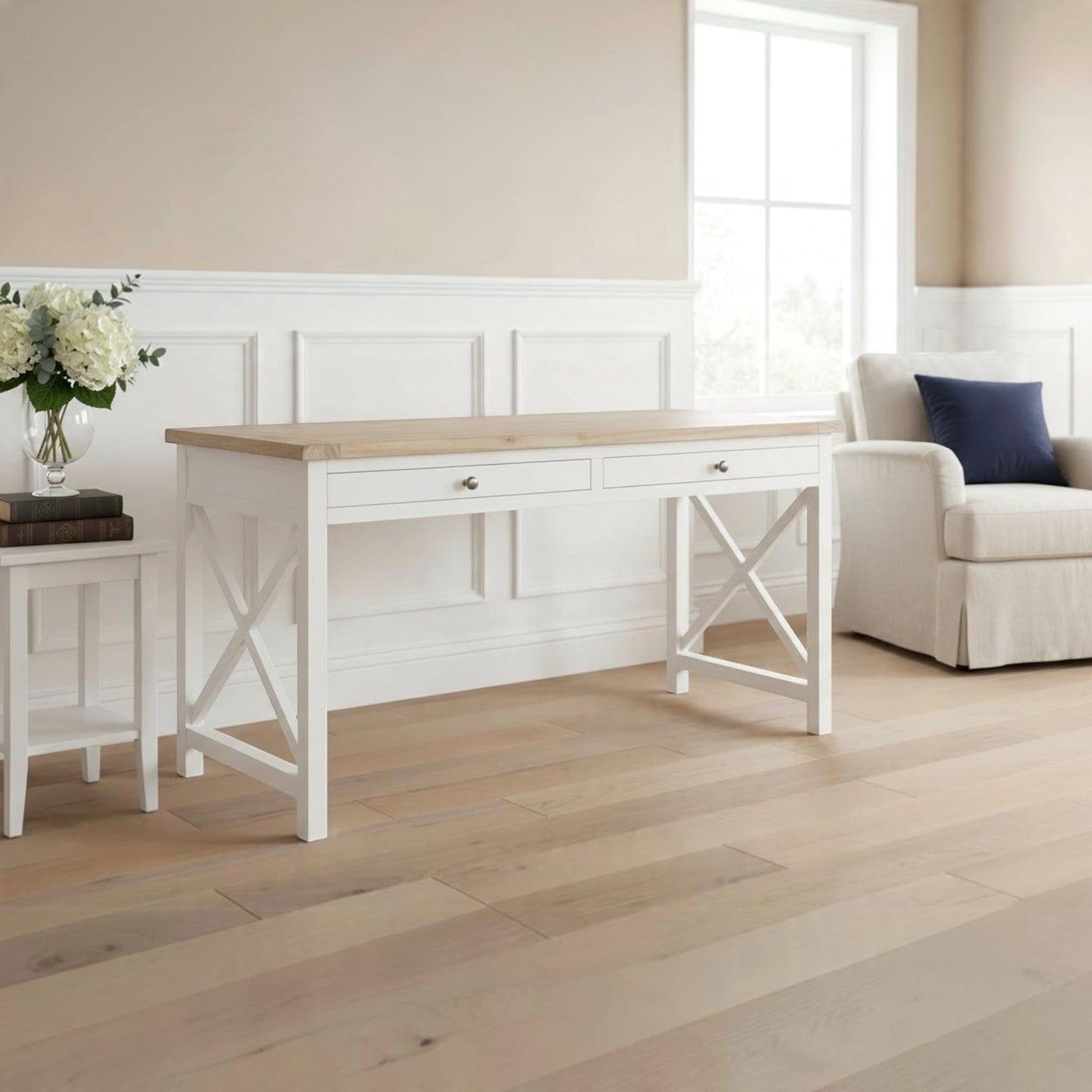 A white writing desk with a light wood top and two drawers, styled in a bright, neutral-toned room next to a small side table with flowers and an armchair.