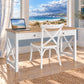 A white writing desk with a light wood top and a matching white chair with a woven seat, set in a bright room with a large window overlooking a beach scene. An open laptop sits on the desk.