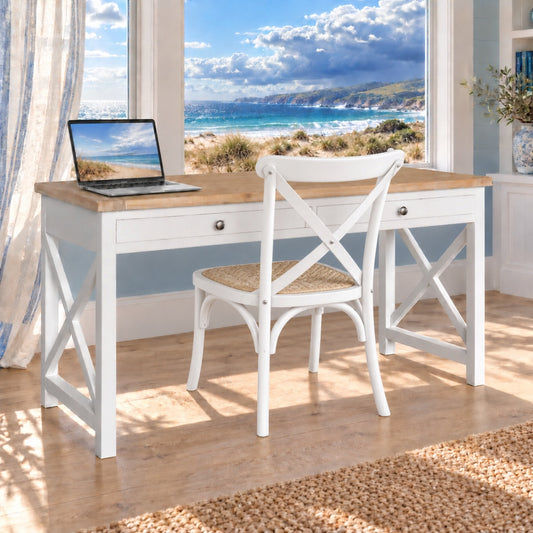 A white writing desk with a light wood top and a matching white chair with a woven seat, set in a bright room with a large window overlooking a beach scene. An open laptop sits on the desk.