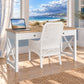 A white desk with a light wood top and matching white wicker chair, set in a bright room with a large window overlooking the ocean.