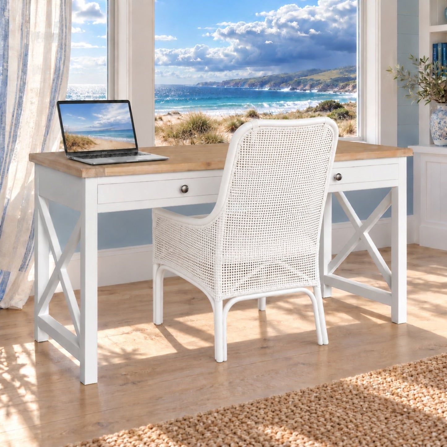 A white desk with a light wood top and matching white wicker chair, set in a bright room with a large window overlooking the ocean.