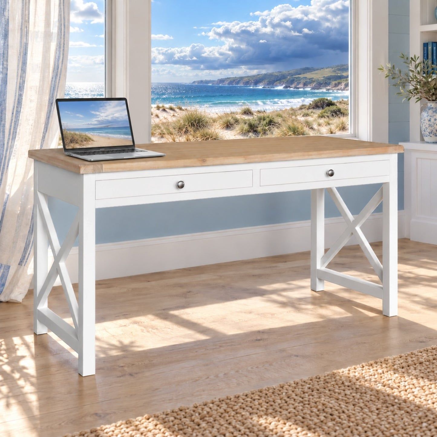 A white writing desk with a light wood top and X-shaped legs, styled in a bright room with a beach view window behind it. An open laptop sits on the desktop.