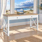 A white writing desk with a light wood top and X-style legs, styled in a bright room with a beach view window and a laptop on top.