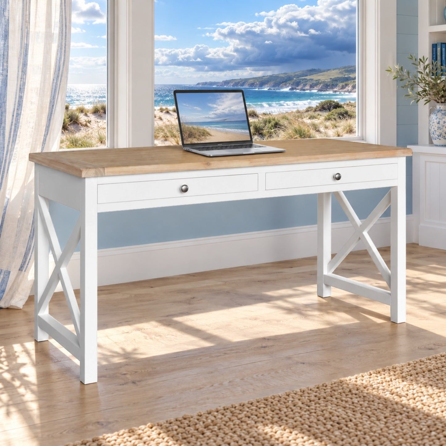 A white writing desk with a light wood top and X-style legs, styled in a bright room with a beach view window and a laptop on top.