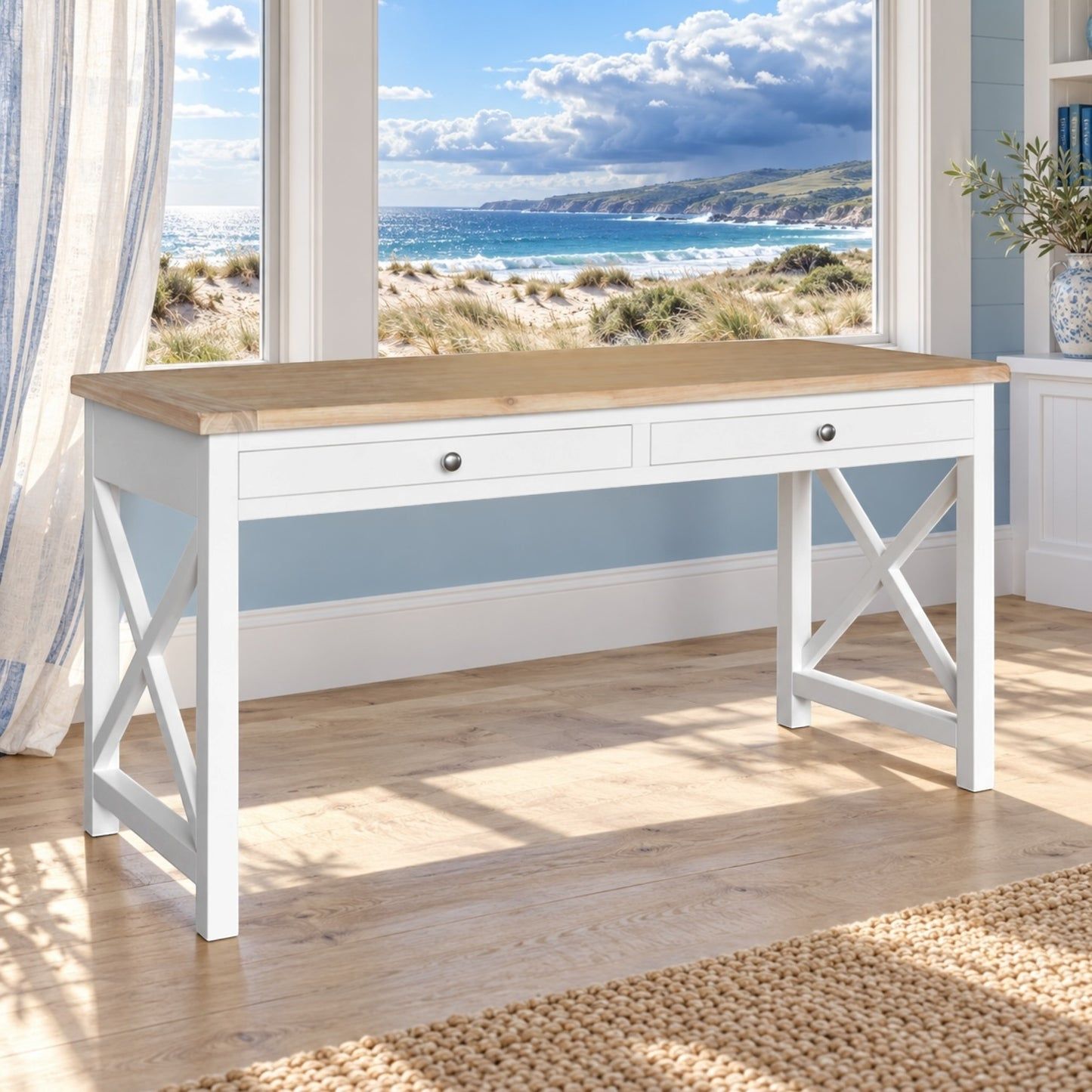 A white writing desk with a light natural wood top and two drawers, positioned in front of a large window overlooking a beach scene. The desk features white X-shaped legs and is set on a light wood floor with a woven rug.