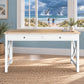 A white writing desk with a light wood top and two drawers, styled in a coastal-themed room with a window showing a beach scene with whales in the background.