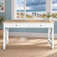 A white writing desk with a light natural wood top, two drawers, and X-shaped legs, styled in a coastal-themed room with a beach window view.