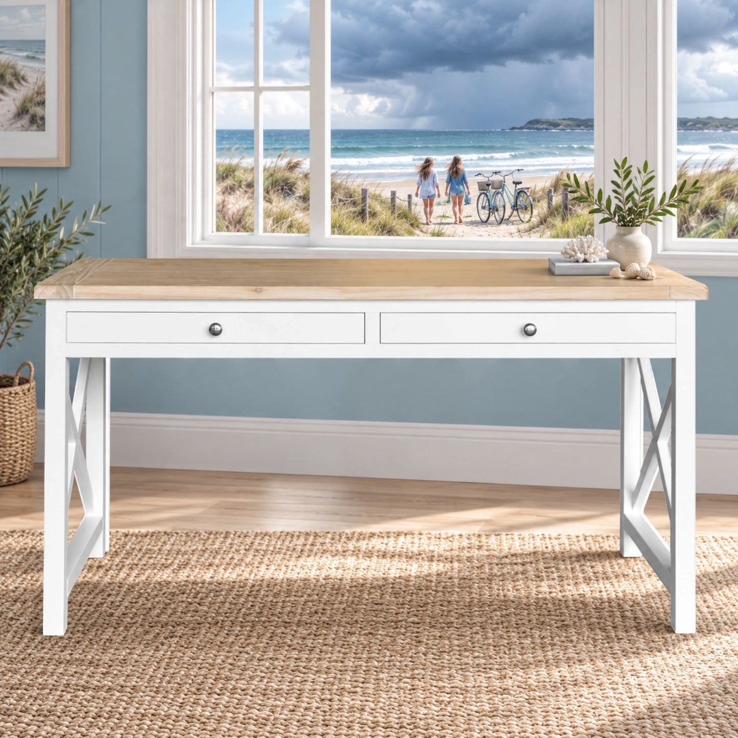 A white writing desk with a light natural wood top, two drawers, and X-shaped legs, styled in a coastal-themed room with a beach window view.