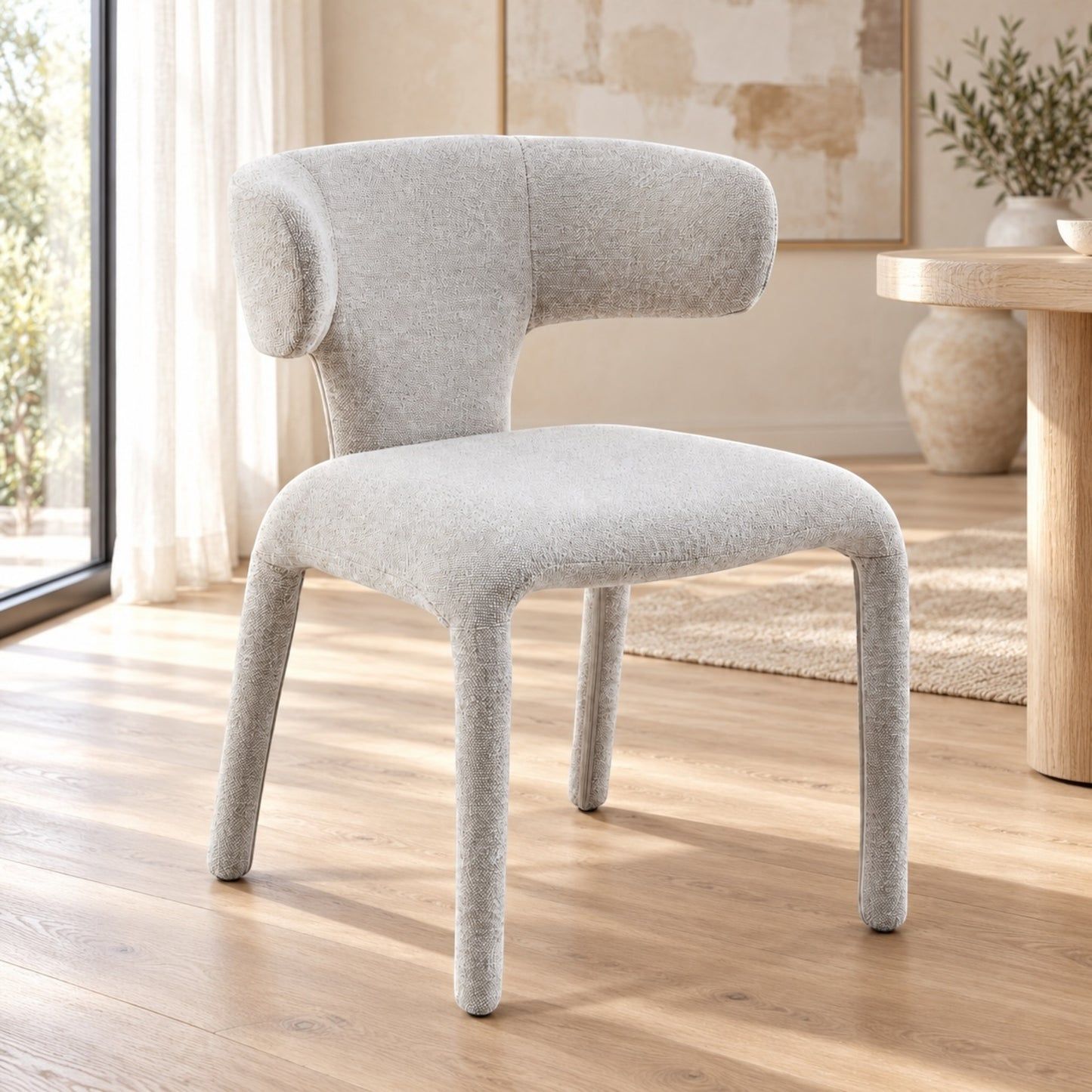 A light gray upholstered accent chair with a curved backrest and tapered legs, shown in a bright, modern living room with wood floors and a window.