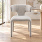 A modern upholstered dining chair with a curved backrest and legs, upholstered in a textured light gray fabric, set in a bright, sunlit room with light wood floors.
