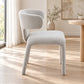 A modern upholstered dining chair with a curved backrest and tapered legs, upholstered in a light gray textured fabric, set in a bright, sunlit room with light wood floors.