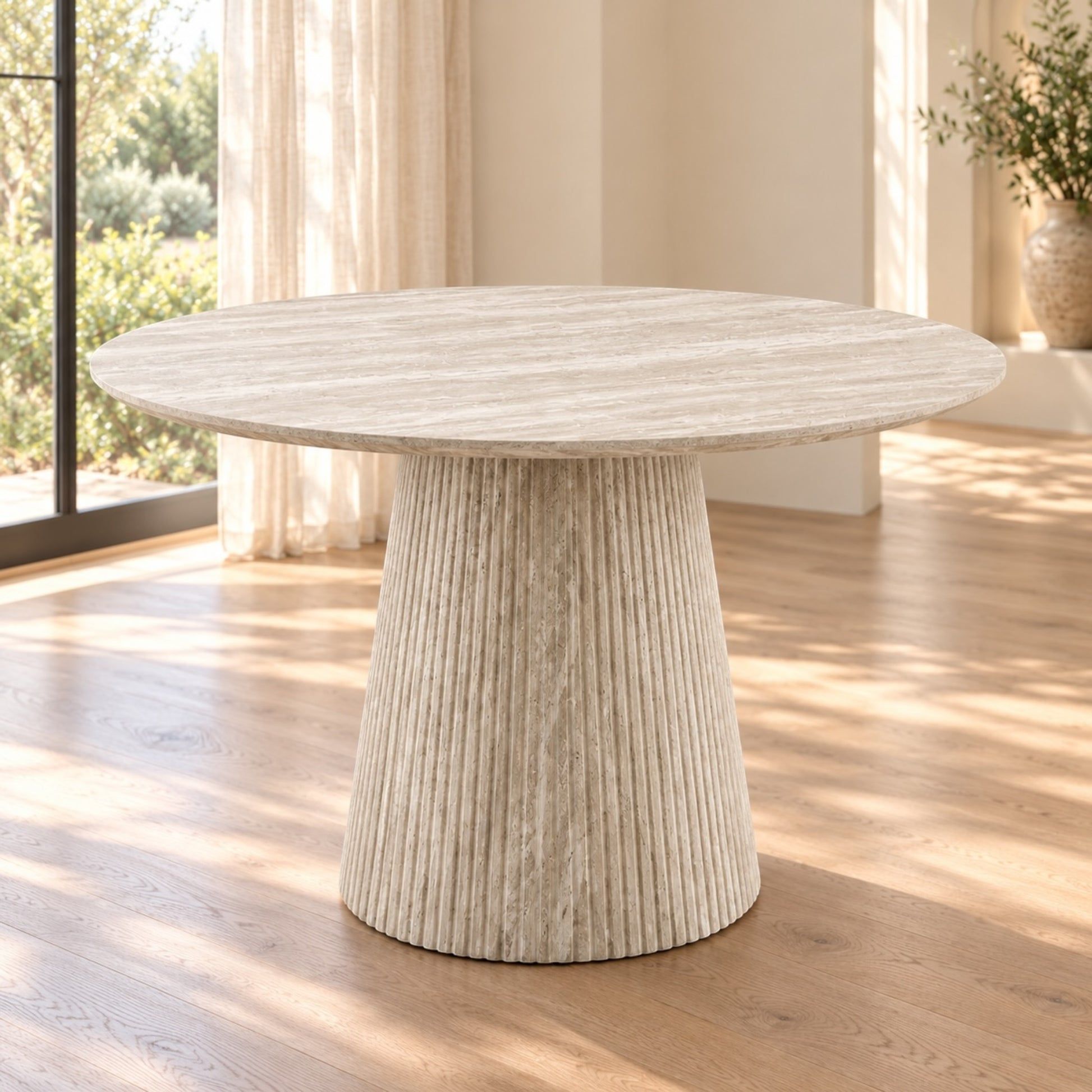 A round dining table with a textured, ribbed pedestal base in a light beige stone or concrete finish, set in a bright room with wooden floors and natural light.