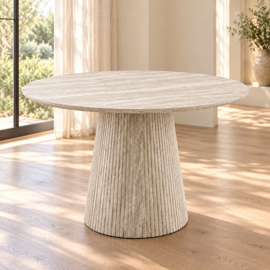 A round dining table with a textured, ribbed pedestal base in a light beige stone or concrete finish, set in a bright room with wooden floors and natural light.