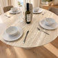 A round dining table with a light beige marble-look surface and matching pedestal base, set with white bowls, plates, wine glasses, cutlery, and a bottle of Uptown Lights wine in a bright dining room.